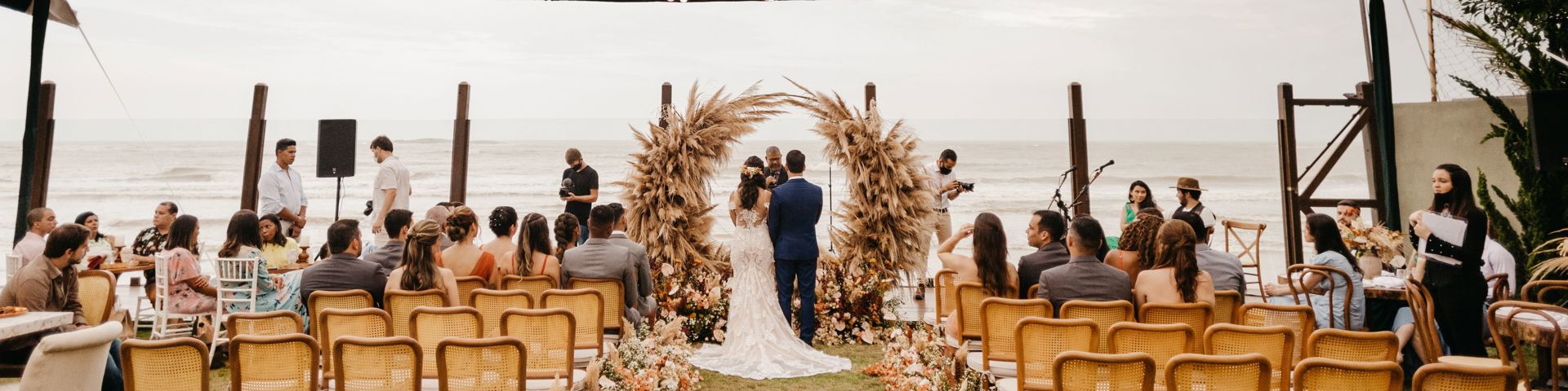Couple exchanging vows under a pampas grass arch in an open-sided marquee by the sea, with guests seated on either side of a flower-lined aisle