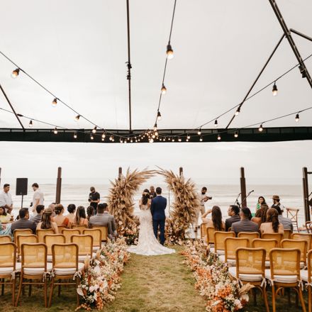 Couple exchanging vows under a pampas grass arch in an open-sided marquee by the sea, with guests seated on either side of a flower-lined aisle
