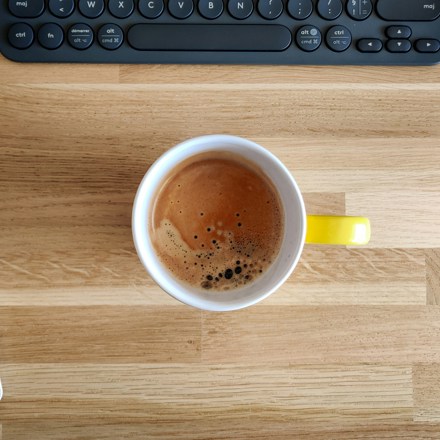 Cup of coffee on a wooden desk beside an open lined notebook and a keyboard