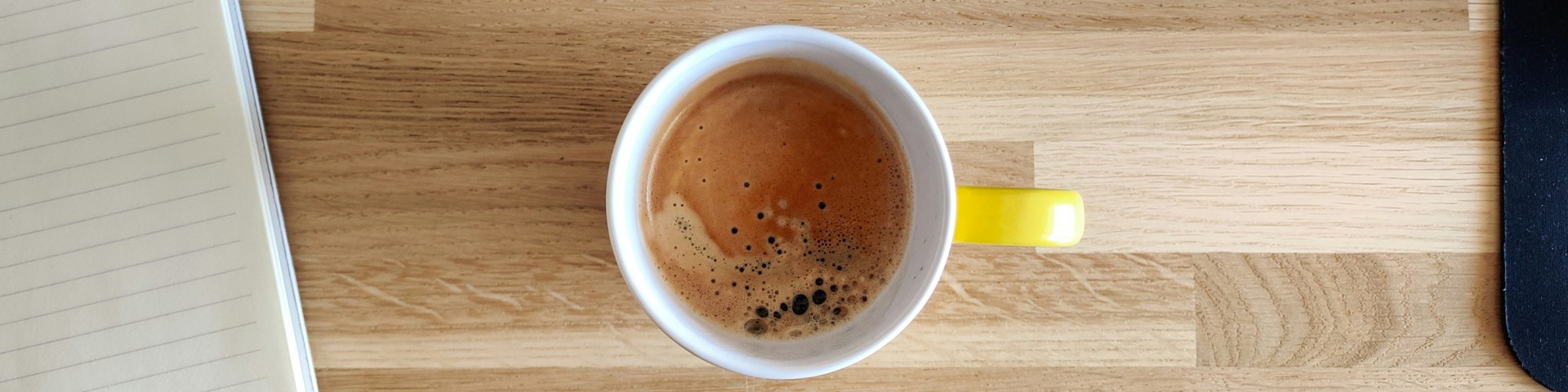 Cup of coffee on a wooden desk beside an open lined notebook and a keyboard