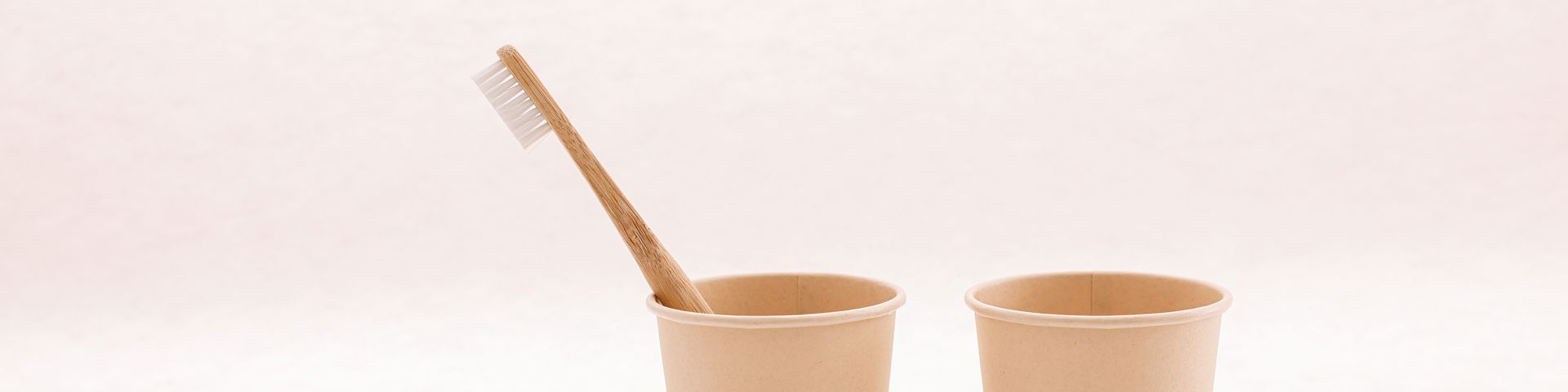 Bamboo toothbrush in a paper cup beside an empty cup on a plain light background
