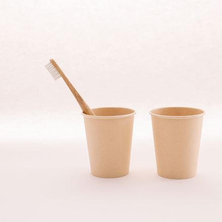 Bamboo toothbrush in a paper cup beside an empty cup on a plain light background