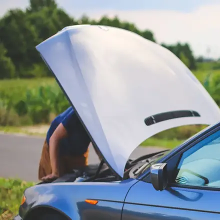 Person checking under the bonnet of a broken-down car on the side of a country road