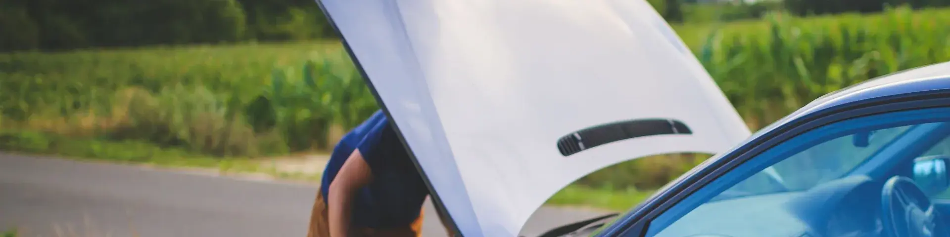 Person checking under the bonnet of a broken-down car on the side of a country road